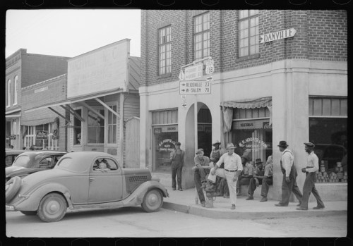 A historic black-and-white photo of a small town street with people gathered outside a store and vintage cars parked nearby.