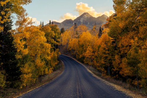 A winding road surrounded by vibrant autumn trees and mountains under a golden sky.