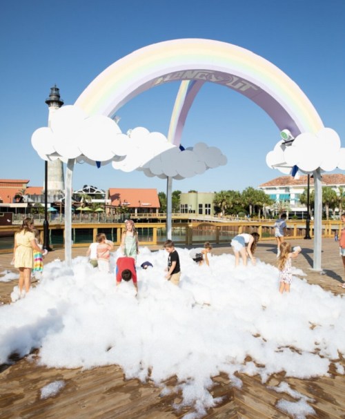 Children playing in a foam pit under a colorful rainbow and clouds at a waterfront location.