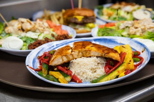 A plate of grilled salmon on rice, surrounded by colorful sautéed peppers, with other dishes in the background.