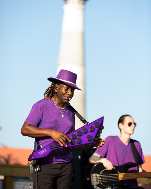 A musician in a purple outfit plays a purple keytar, while another musician on bass stands nearby, both performing outdoors.