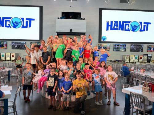 A large group of children in colorful shirts poses together on a stage at The Hangout, with screens in the background.