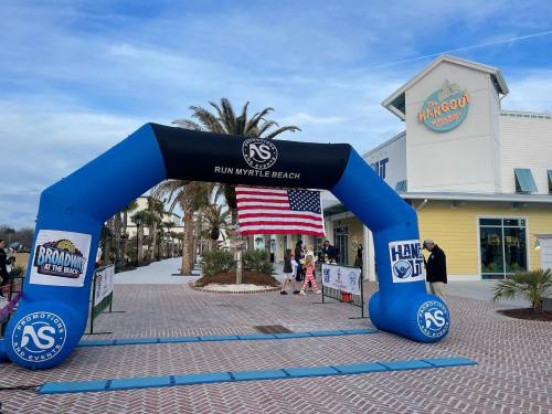 An inflatable arch with "Run Myrtle Beach" and an American flag, set near a beachside venue with palm trees.