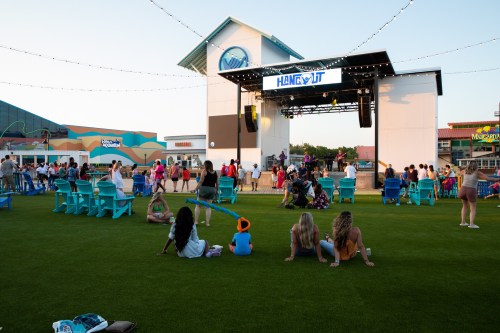 A lively outdoor event with a stage, people seated on grass, and colorful buildings in the background during sunset.