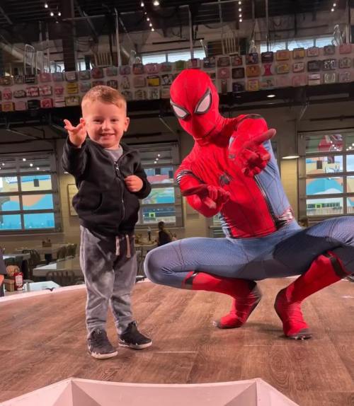 A young boy poses with Spider-Man, both making playful hand gestures in a lively indoor setting.