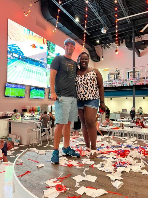 A man and woman stand on a stage covered in confetti, smiling, with a large screen displaying a sports event in the background.