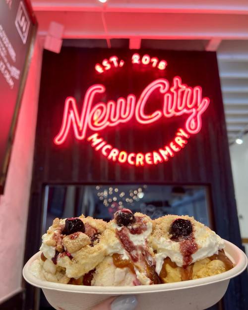 A hand holds a bowl of ice cream topped with berries and crumbs in front of a neon sign that reads "New City Microcreamery."