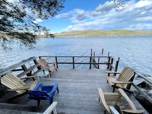 A wooden deck with Adirondack chairs overlooks a calm lake surrounded by trees and mountains under a blue sky.