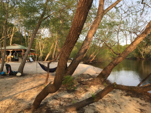 A serene riverside scene with sandy shore, trees, a hammock, and a small shelter in the background.