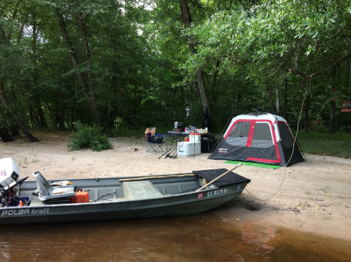 A small boat on the water near a sandy shore with a tent and camping setup in a wooded area.