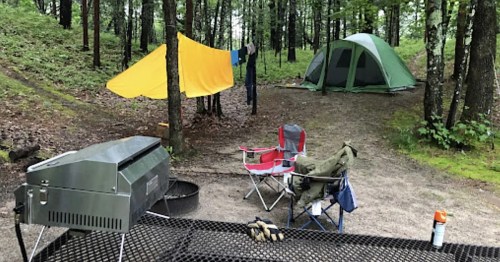 A campsite with a grill, chairs, a yellow tarp, and a green tent surrounded by trees.