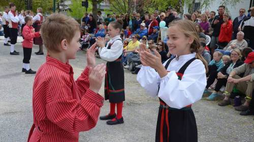 Two children in traditional costumes clap and dance together, while a crowd watches in the background.