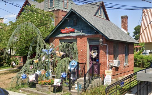 A charming brick house with a sloped roof, colorful decorations, and a whimsical blue character in the front yard.