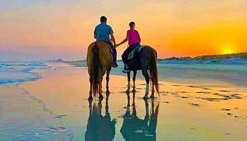 A couple rides horses along a beach at sunset, their reflections visible in the wet sand.