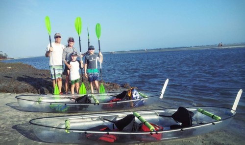 A family stands on the shore with kayaks, holding paddles, against a scenic water backdrop.
