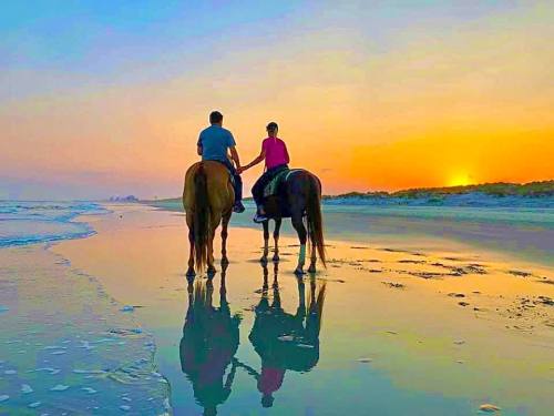 A couple rides horses along a beach at sunset, their reflections visible in the wet sand.