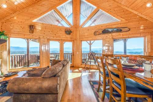 Cozy wooden cabin interior with large windows, offering mountain views, a comfortable sofa, and a dining area.