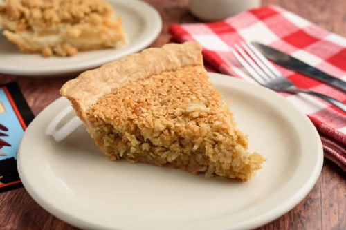 A slice of golden brown pie on a white plate, with a crumbly topping, set on a wooden table with a checkered napkin.