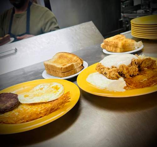 Two yellow plates with breakfast items: eggs, hash browns, sausage, fried chicken, and toast in a diner setting.