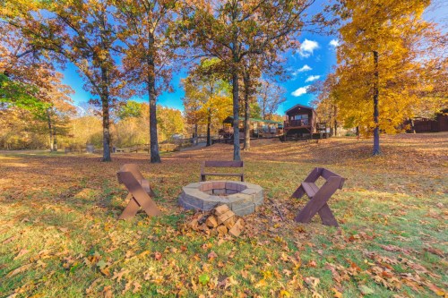 A cozy fire pit surrounded by wooden benches, set in a colorful autumn landscape with trees and a cabin in the background.