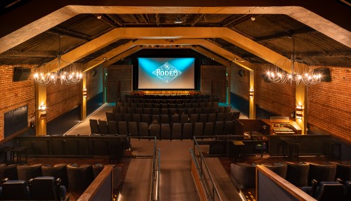 Interior view of a theater with a large screen, rows of seats, and chandeliers, showcasing a cozy atmosphere.