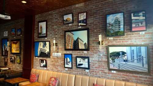 A brick wall adorned with various framed paintings of buildings, above a cozy seating area with decorative pillows.