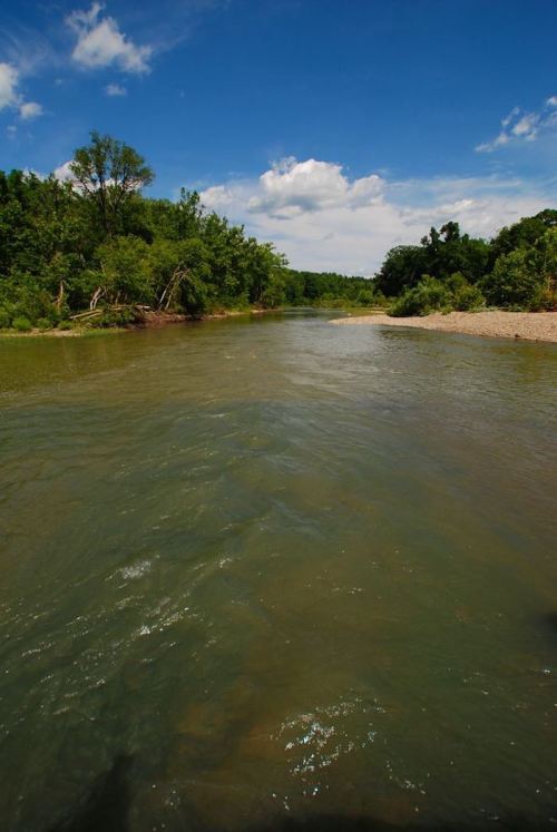 A serene river flows through lush greenery under a bright blue sky with scattered clouds.