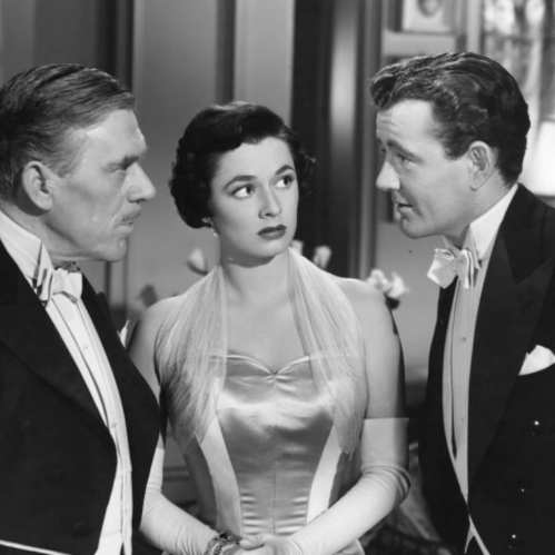 A black-and-white scene featuring three elegantly dressed individuals in formal attire, engaged in conversation.