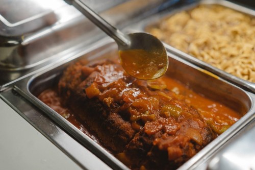 A serving spoon ladles sauce over a meatloaf in a metal tray, with another tray of pasta in the background.