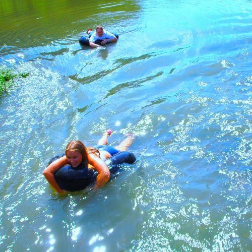 Two people swim in a sparkling blue river, one holding a float while the other swims nearby.