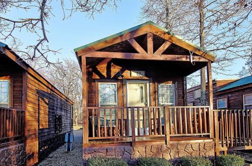 A cozy wooden cabin with a porch, surrounded by trees and other similar cabins in a serene outdoor setting.