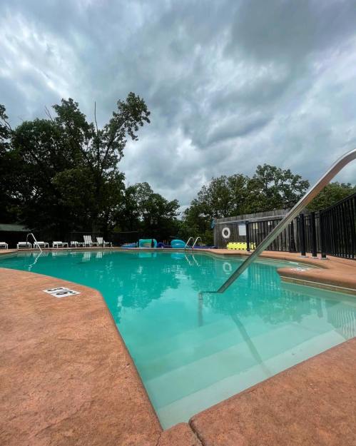 A swimming pool with clear water, surrounded by trees and a cloudy sky, featuring a ladder and lounge chairs.