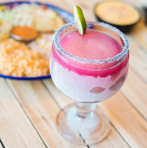 A colorful margarita with a lime wedge, served in a glass, with a plate of food in the background.