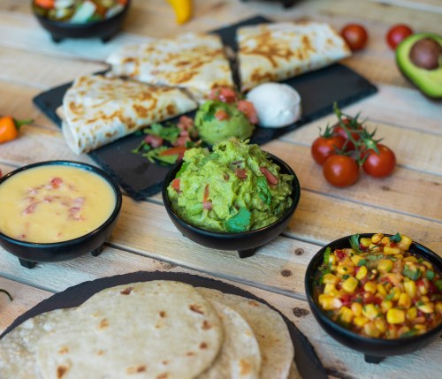 A colorful spread of tortillas, guacamole, cheese dip, corn salad, and fresh tomatoes on a wooden table.