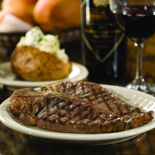 A grilled steak on a plate, accompanied by a baked potato and a glass of red wine in the background.