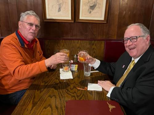 Two men clink glasses in a cozy restaurant, smiling at the camera with drinks in hand.