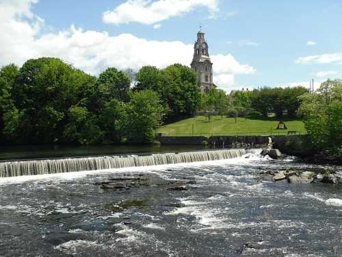 A serene river with a waterfall, surrounded by lush greenery and a historic building in the background under a blue sky.