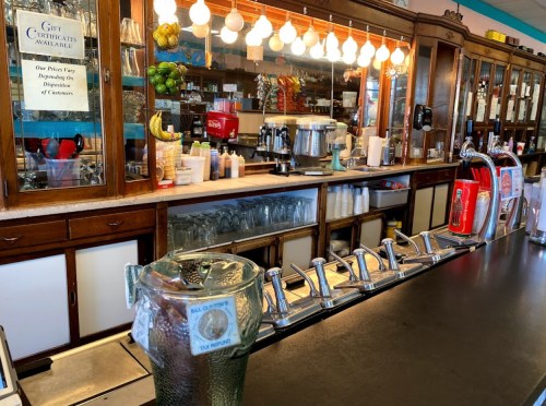 A vintage diner counter with a soda fountain, glassware, and decorative fruit in the background.