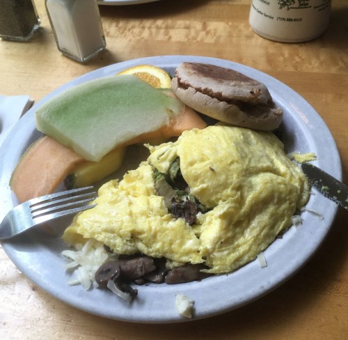 A plate with an omelet, sautéed mushrooms, shredded cheese, and slices of cantaloupe, honeydew, and an English muffin.