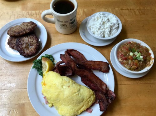 A hearty breakfast plate with an omelet, bacon, sausage, cottage cheese, and a side of fruit, accompanied by coffee.