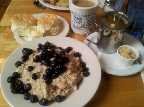 A plate of oatmeal topped with blueberries, served with biscuits, butter, coffee, and a small container of sugar.
