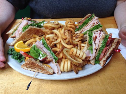 A plate of a club sandwich with turkey, lettuce, and tomato, served with curly fries and a slice of orange.