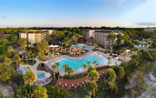 Aerial view of a resort with a large pool, lounge chairs, and surrounding greenery under a clear sky.