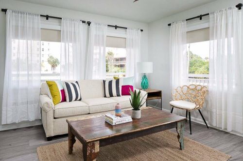 Bright living room with a beige sofa, colorful pillows, a wooden coffee table, and large windows with sheer curtains.