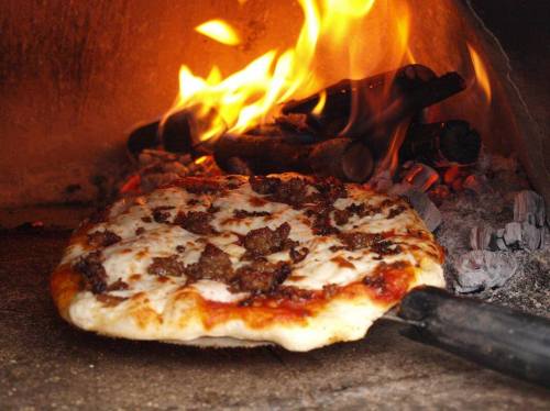 A freshly baked pizza with melted cheese and toppings, being pulled from a wood-fired oven with flames in the background.