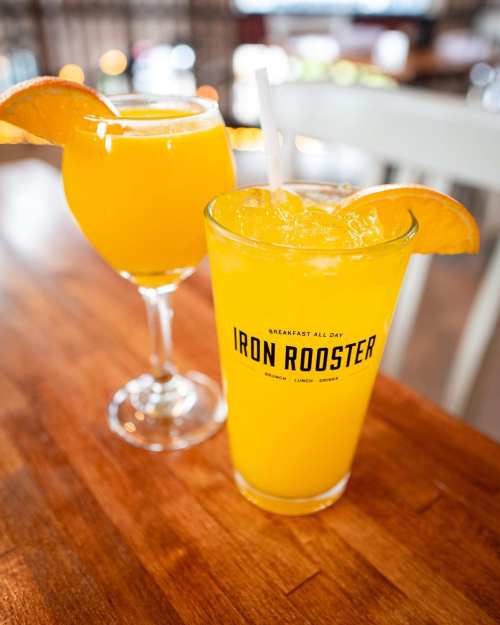 Two orange drinks on a wooden table, one in a tall glass and the other in a stemmed glass, both garnished with orange slices.