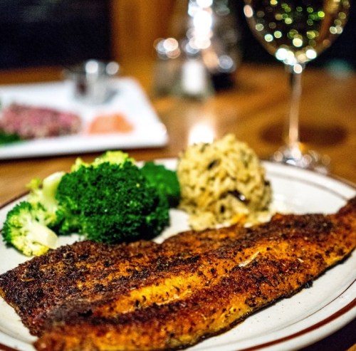 A plate of seasoned fish fillet with broccoli and rice, accompanied by a glass of white wine in a cozy dining setting.