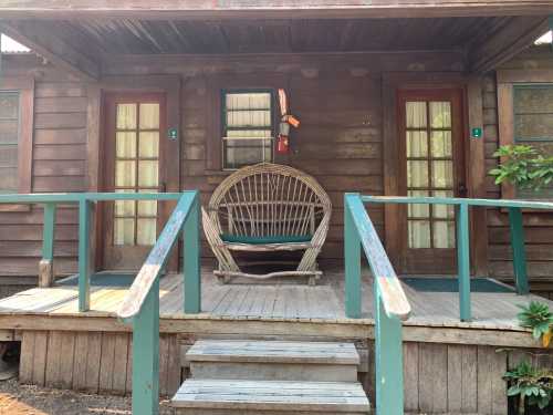 A rustic wooden cabin with two doors and a large wicker chair on the porch, surrounded by greenery.