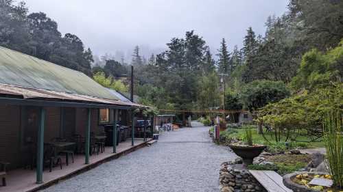 A gravel path leads through a serene garden with trees, a building on the left, and misty mountains in the background.