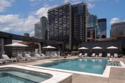 Rooftop pool area with lounge chairs and umbrellas, surrounded by modern skyscrapers under a partly cloudy sky.